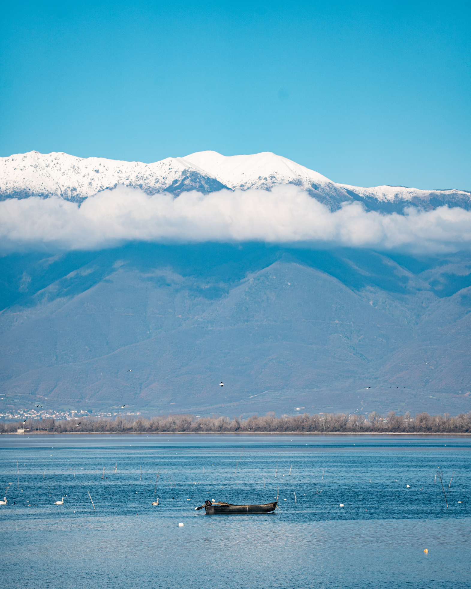 A small boat on Lake Kerkini beneath snow-covered mountains and a band of low cloud.