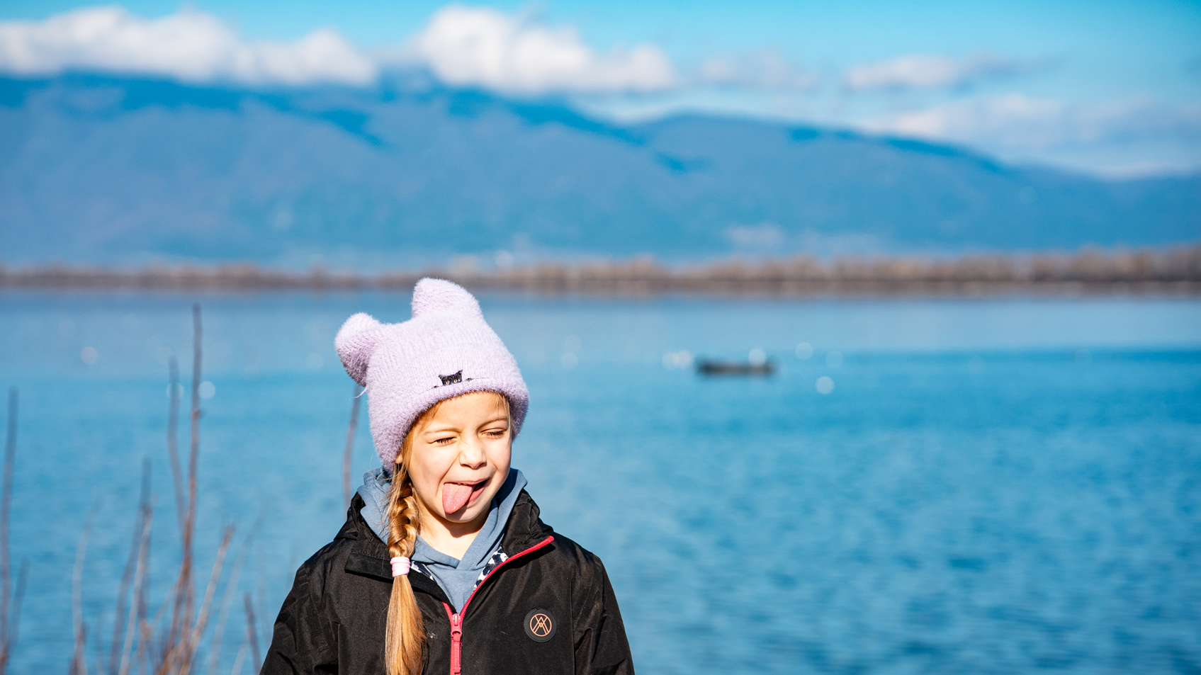 Melina sticking her tongue out in a pink woolly hat beside Lake Kerkini, with snowy mountains behind her.