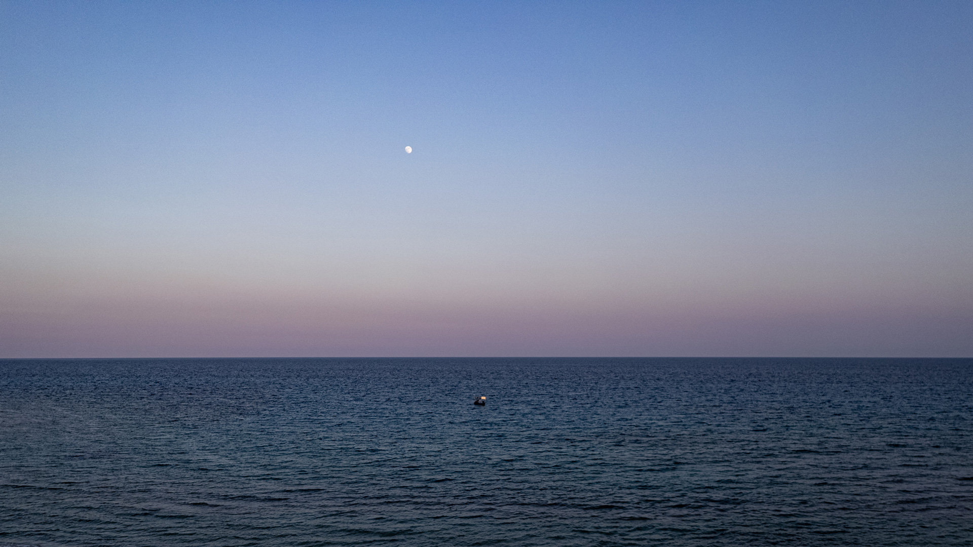 The moon above a calm sea beneath a soft blue and pink twilight sky in Chalkidiki.
