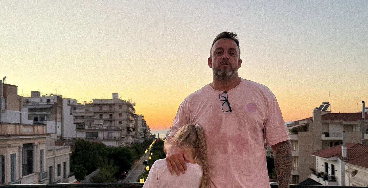 Paul standing above the rooftops of Patras in a pink T-shirt at sunset.
