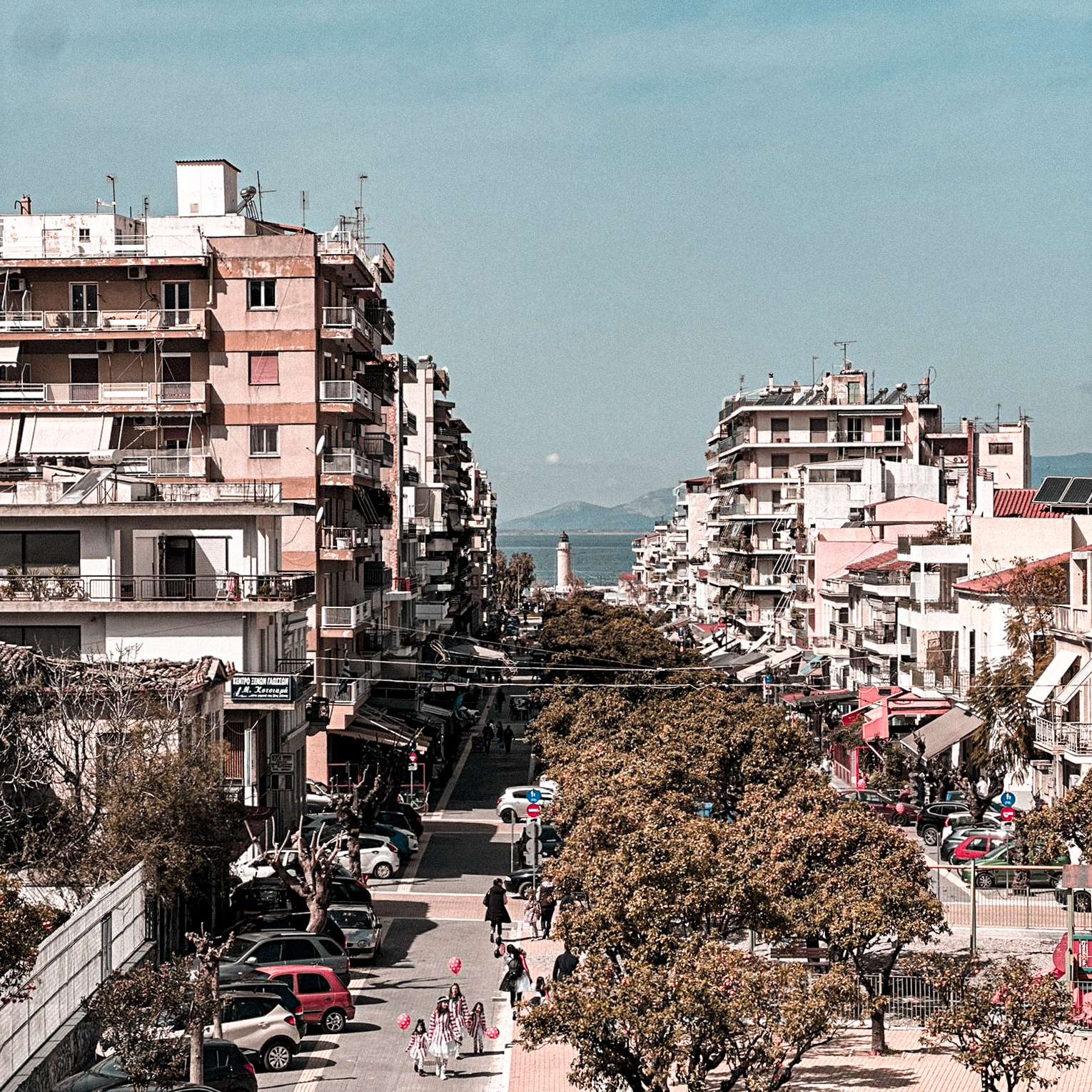 View down a sunlit street in Patras, with apartment balconies, trees, and mountains in the distance.