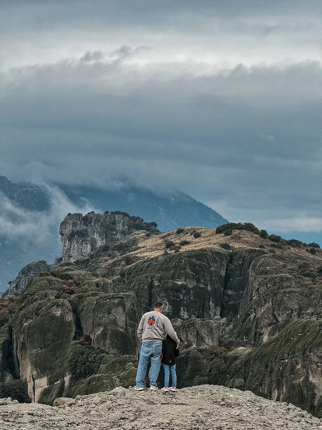 Two people standing together on a rocky viewpoint overlooking Meteora beneath dark clouds.