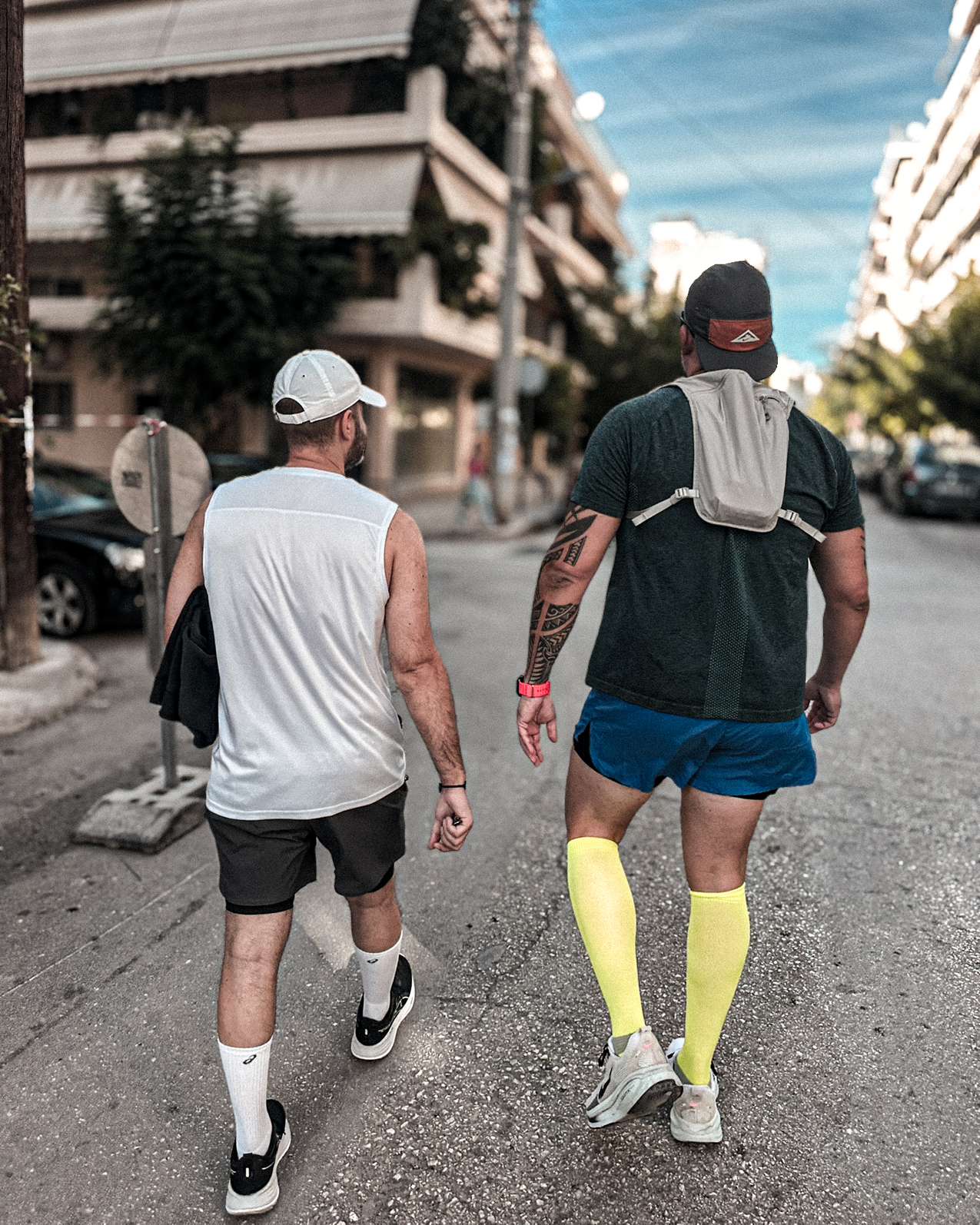 Paul and Iasonas seen from behind on a Patras street, wearing running clothes with towels over their shoulders.