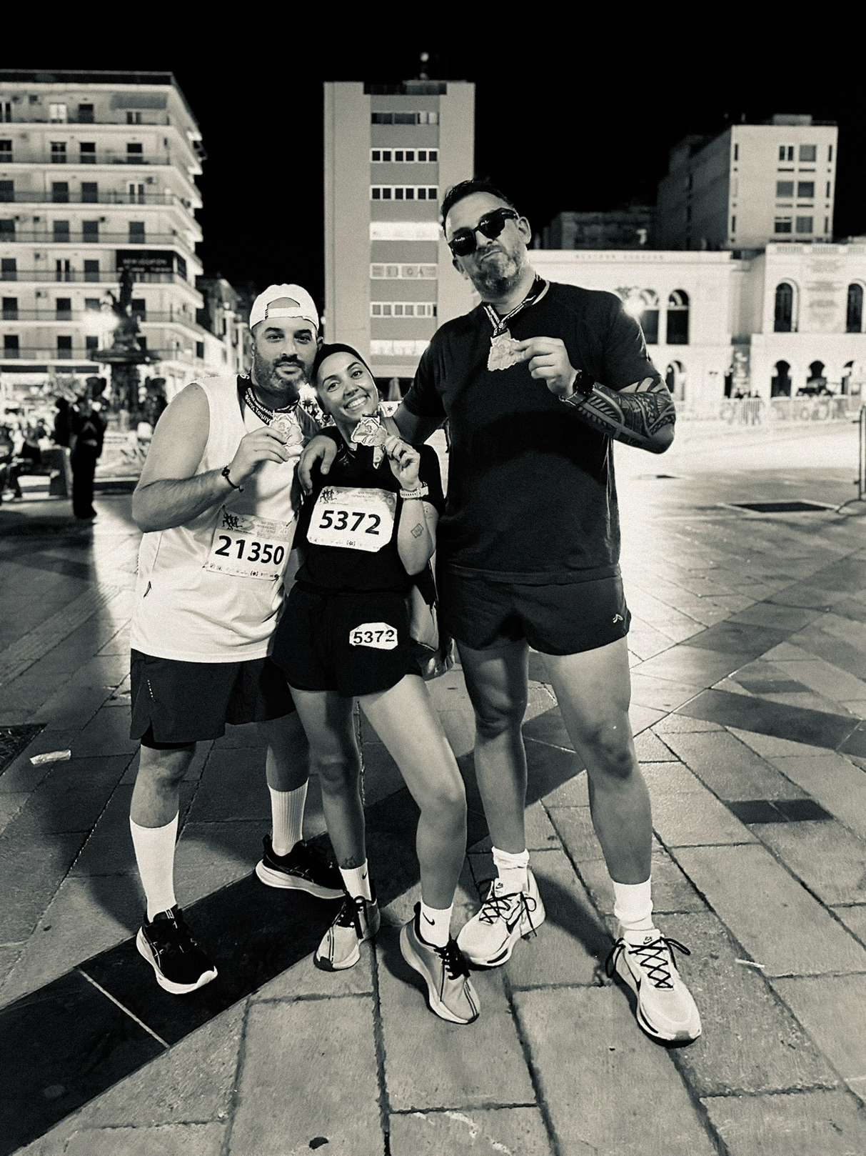 Black-and-white photograph of three friends posing with race medals in a Patras square at night.