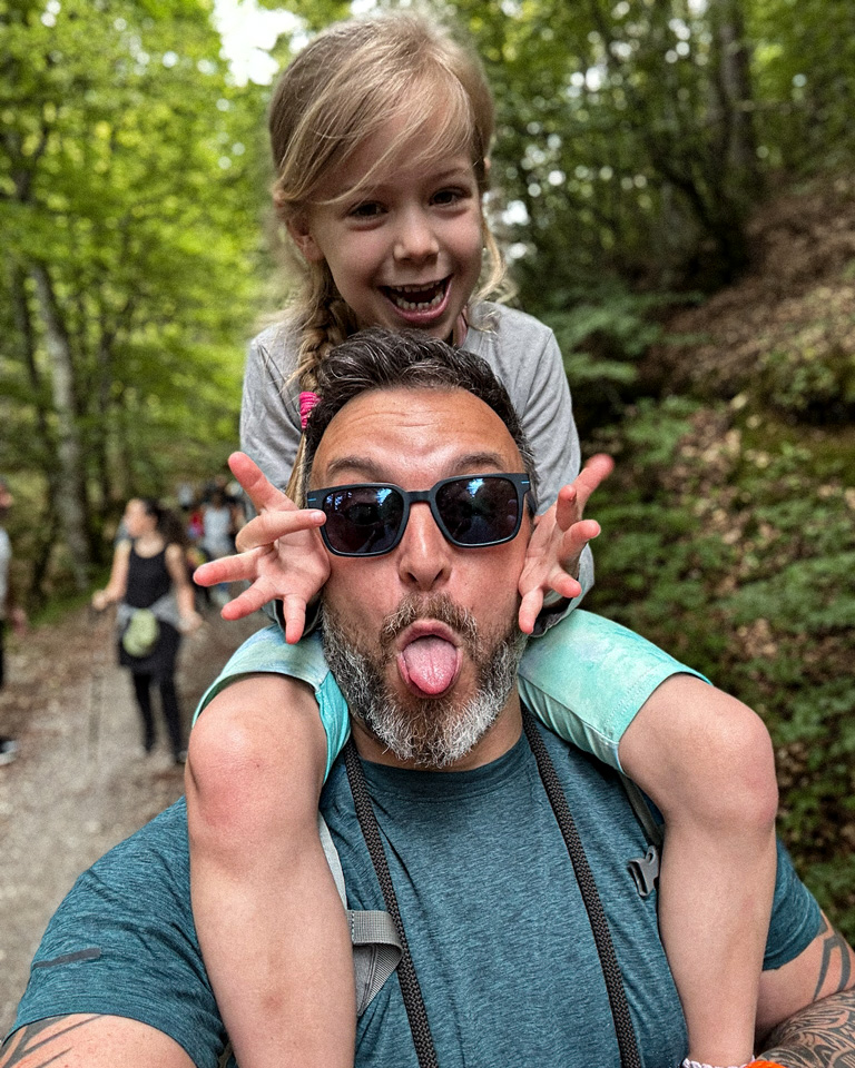 Paul carrying Melina on his shoulders in the forest, both making playful faces for the camera.