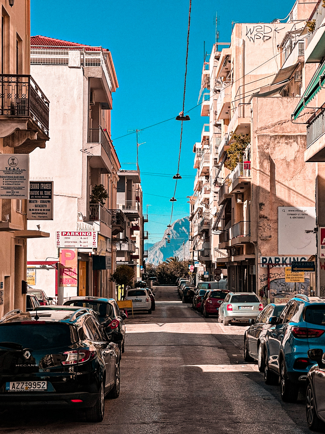 Sunlit street in Patras lined with parked cars and apartment buildings, with a mountain in the distance.