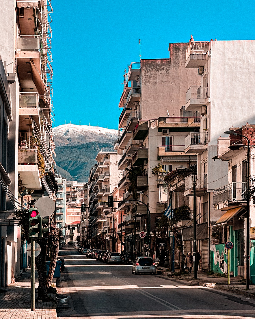 Street in Patras framed by apartment buildings, with a snow-dusted mountain in the distance.