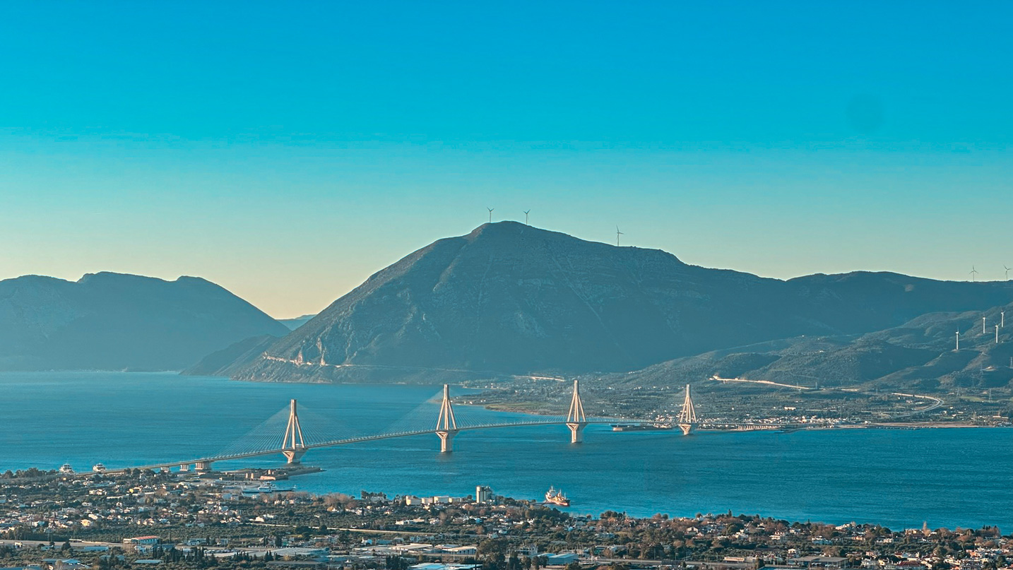 Rio–Antirrio Bridge spanning blue water beneath mountains and hillside wind turbines.