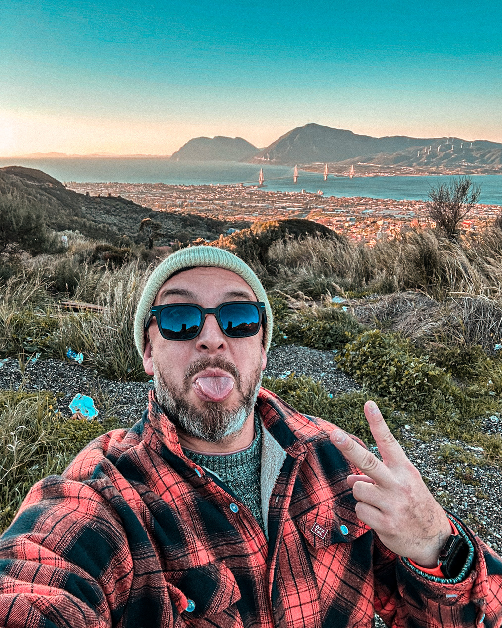 Self-portrait making a peace sign above Patras, with the Rio–Antirrio Bridge and mountains behind.