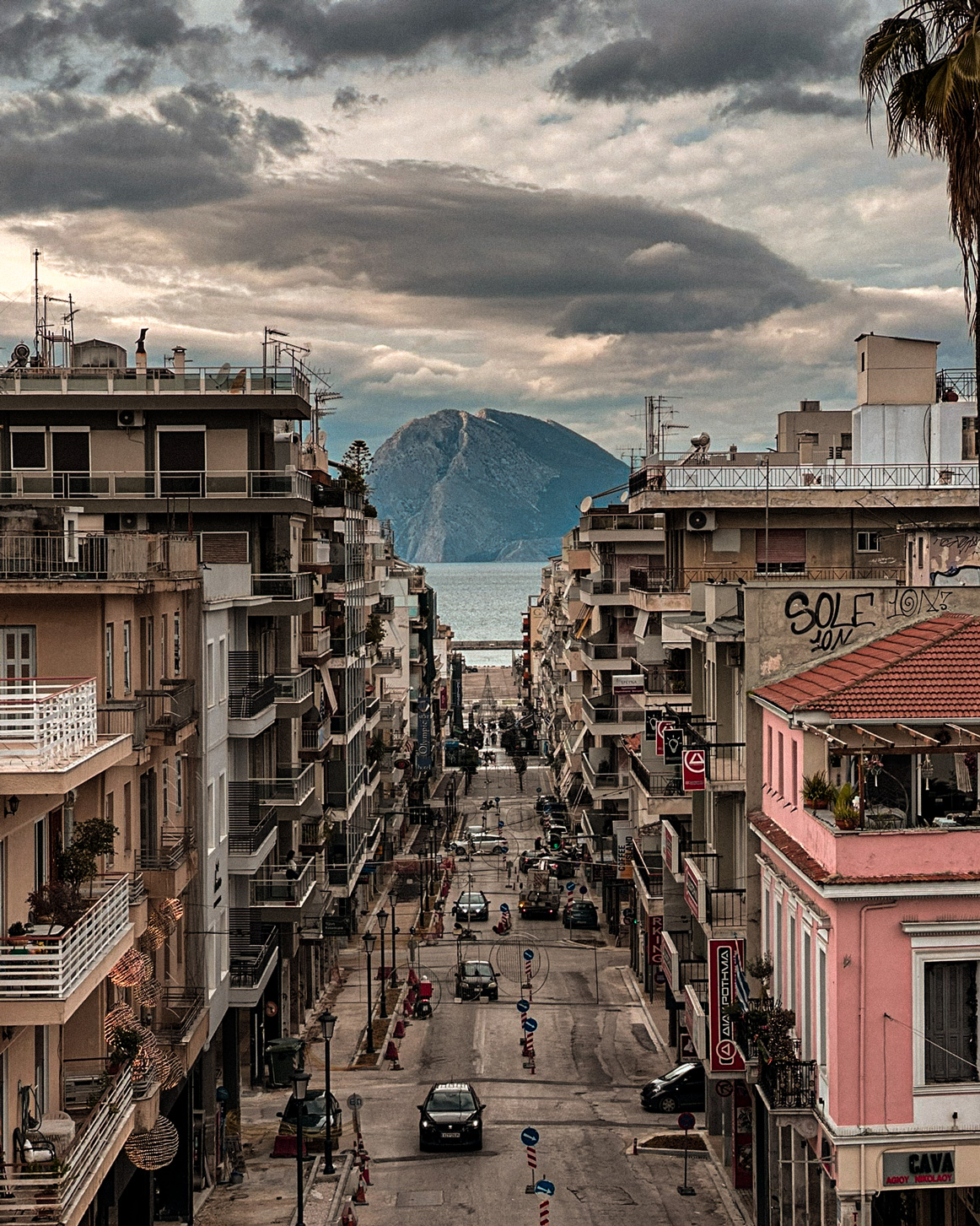 View down a Patras street toward the sea and distant mountains under a cloudy sky.