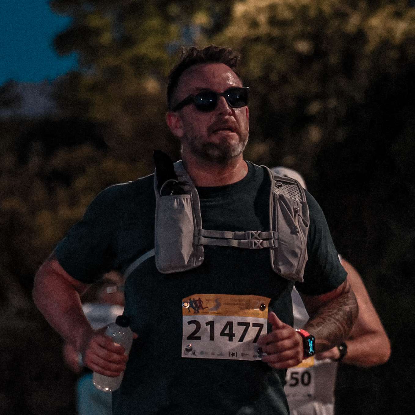 Close-up of Paul running the Patras half marathon in sunglasses and a hydration vest, wearing bib 21477.