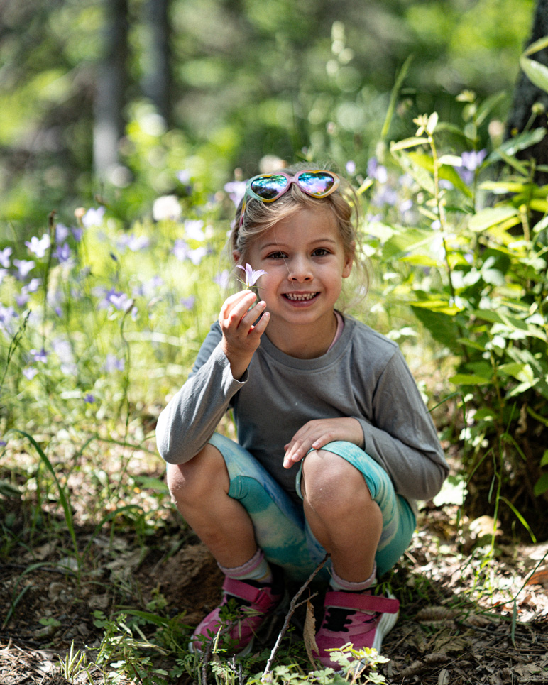 Melina crouching among plants and white flowers on a sunlit forest slope.