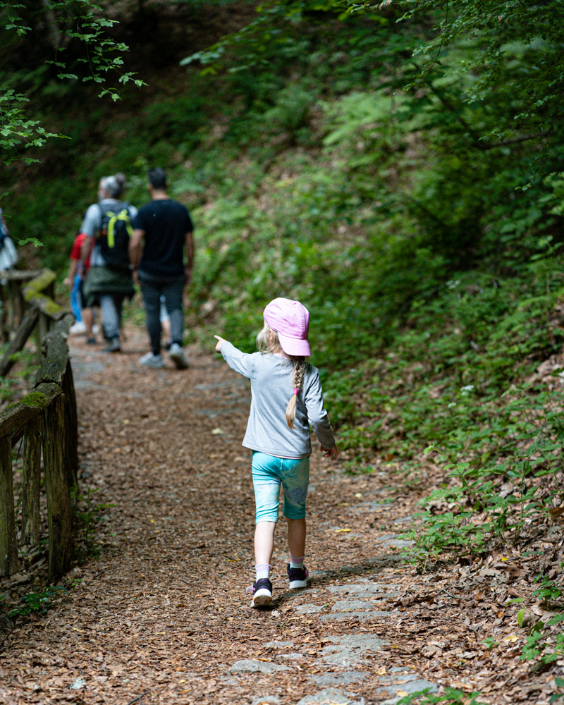 Melina in a pink hat walking along a forest trail behind a group of adults in Ano Milia.