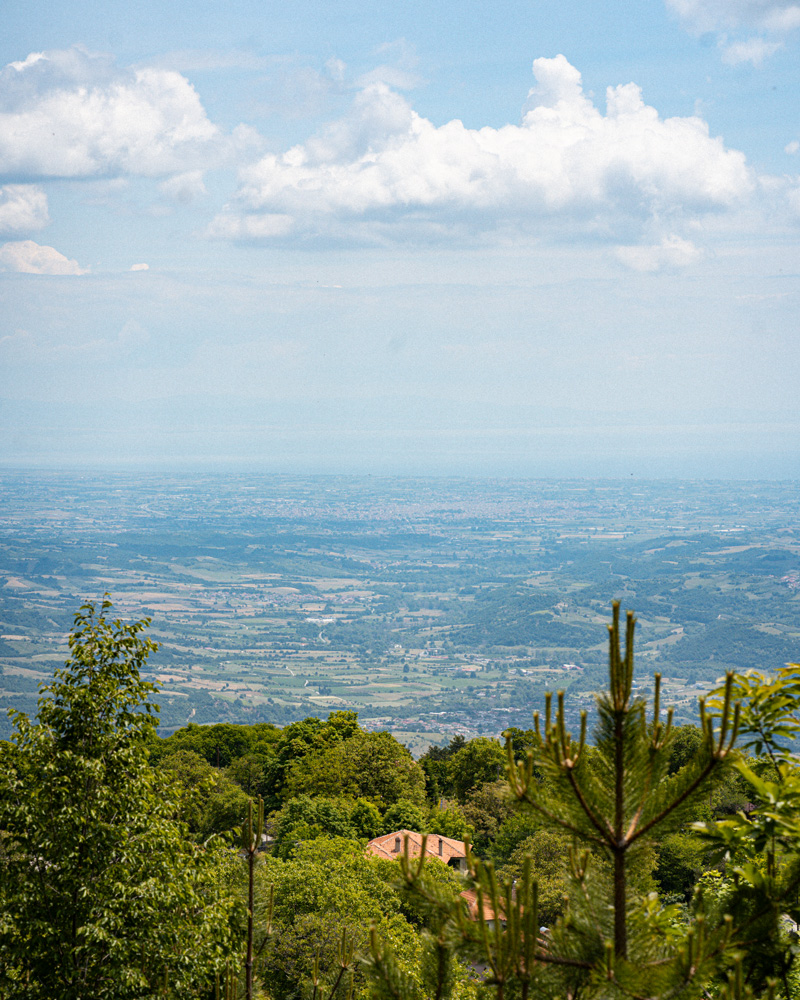 View from the wooded slopes of Ano Milia across a broad plain beneath a partly cloudy sky.