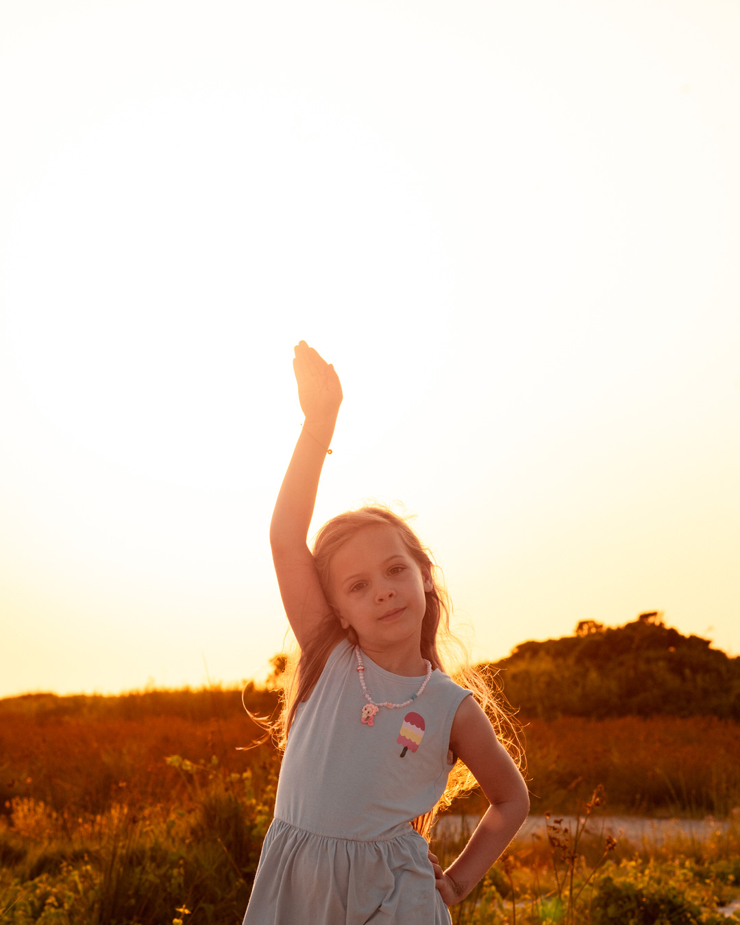 Melina raising her hand against the sunlight, surrounded by the warm orange glow of sunset.