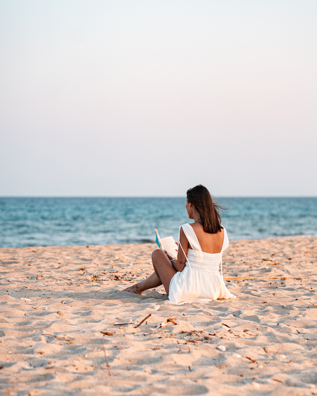 Eirini in a white dress sitting on the beach, with softly lit sand and sea around her.