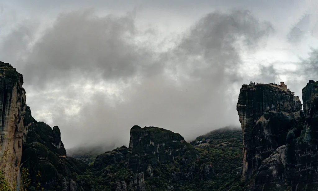 Panoramic view of a clifftop monastery and towering Meteora rocks beneath low, heavy clouds.