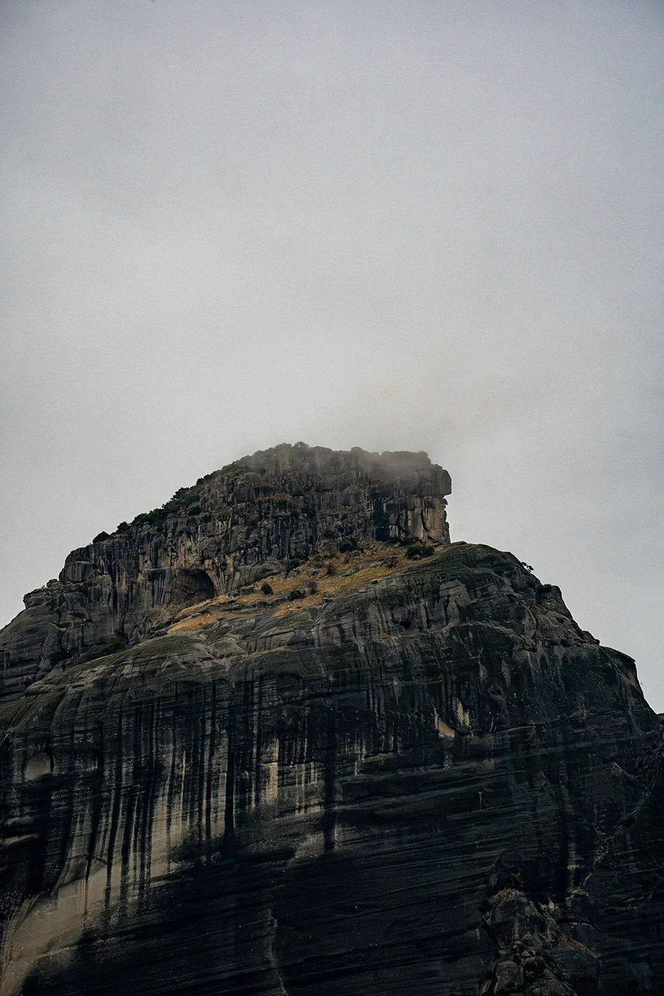 Dark, layered rock formation at Meteora with its summit partly hidden by low cloud.