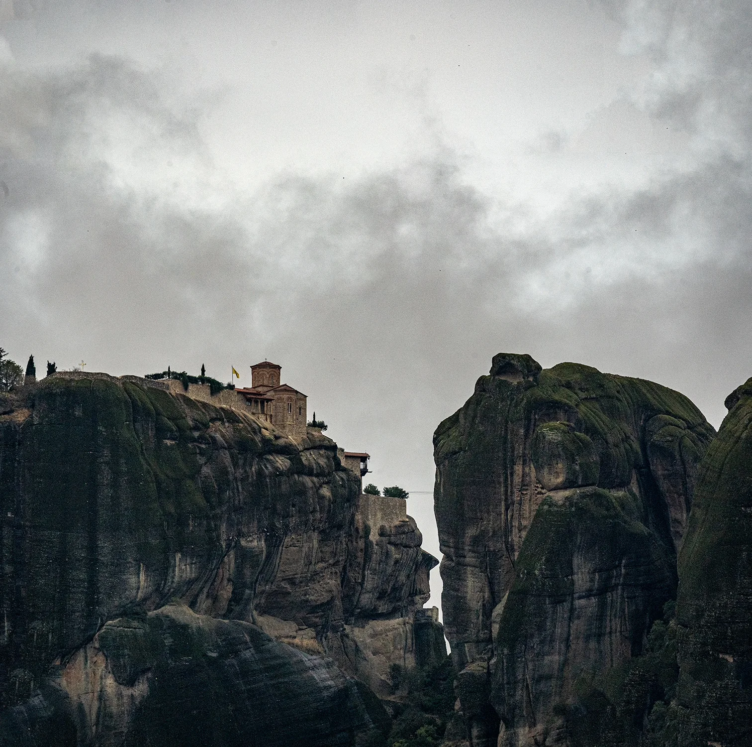 Monastery on a sheer cliff beside towering rock formations at Meteora beneath an overcast sky.