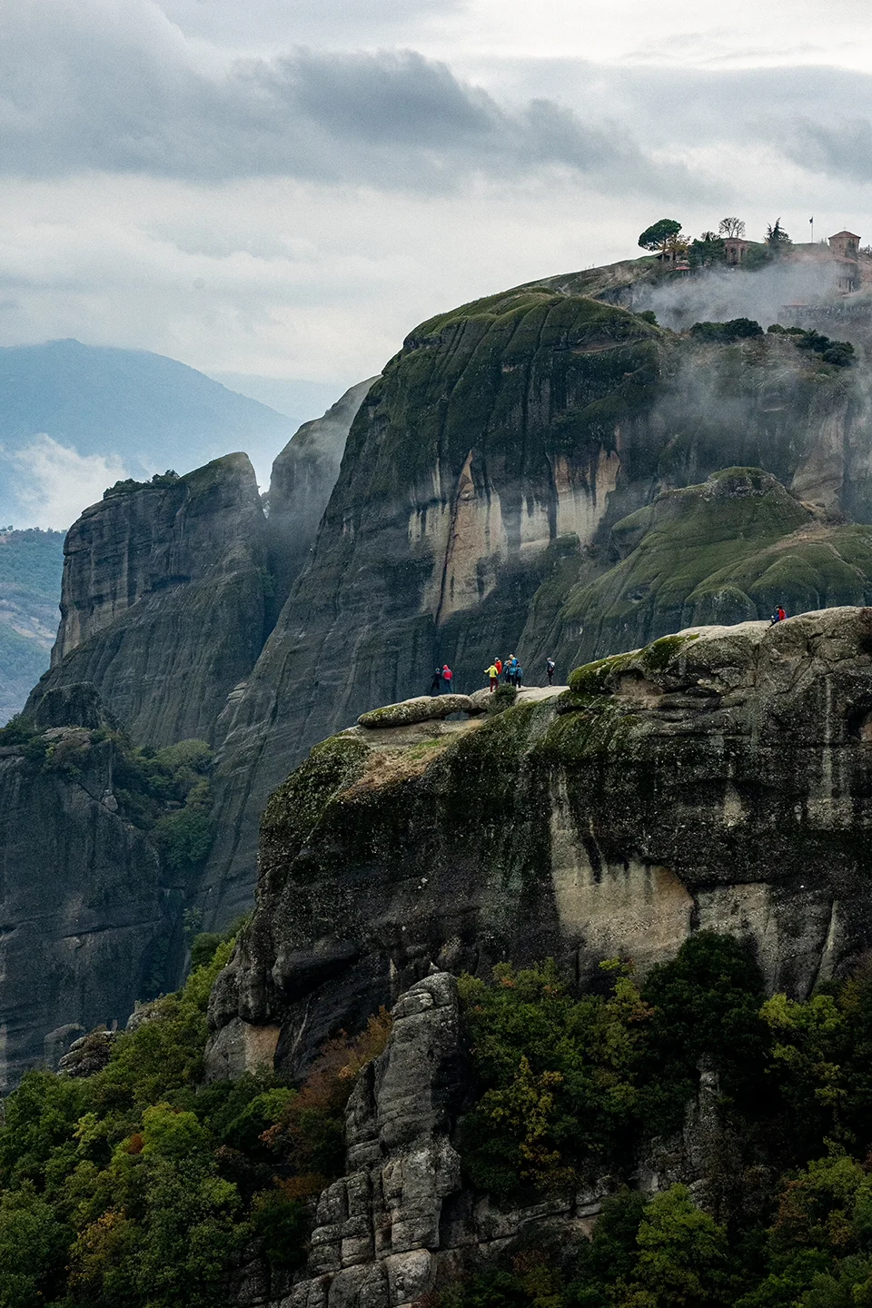 Low cloud drifting between steep, layered rock formations and wooded slopes at Meteora.
