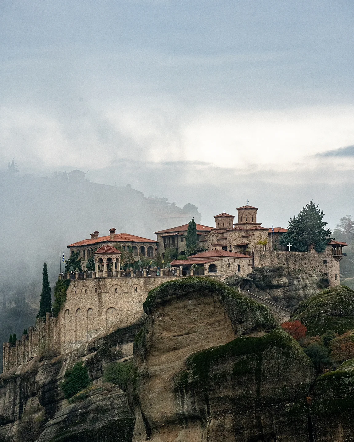 Monastery buildings lining a sheer cliff at Meteora, with mist drifting across the rocks behind.